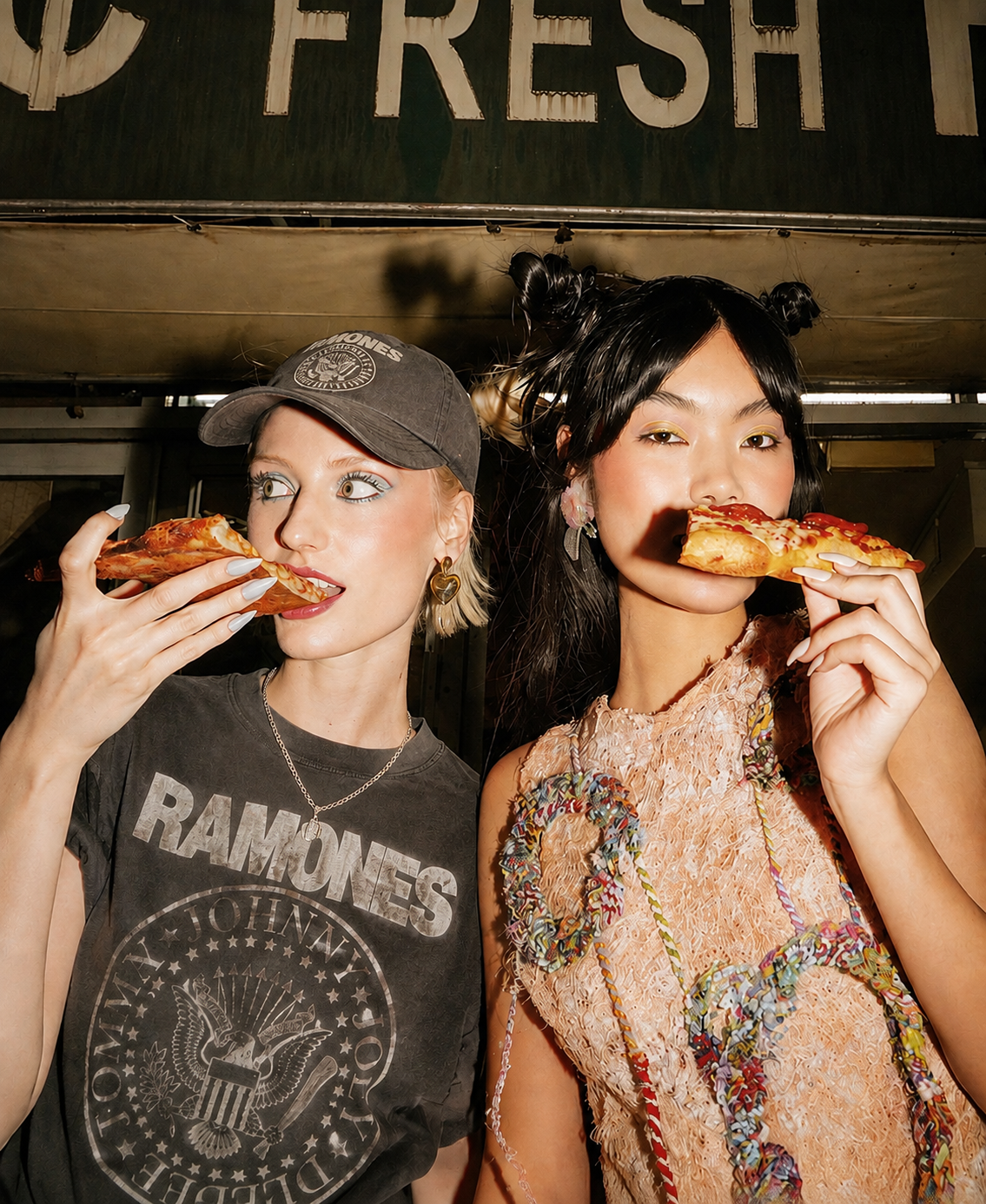 Två vänner med pizzaslices i band-tee och spetsklänning